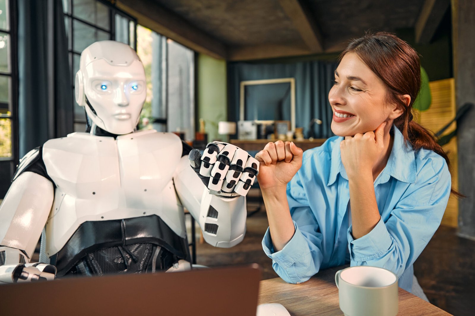 Young woman with a cup of coffee and a humanoid robot working while sitting at a laptop in a modern office, fist to fist gesture. Collaboration between humans and artificial intelligence.  AI Marketing